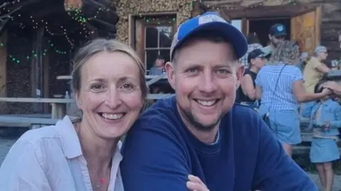 Caroline Stafford Head and shoulders image of Caroline in a light shirt and Gareth in a dark blue sweatshirt. They are both smiling. Behind them you can see a small cluster of people including a child.