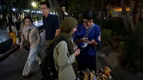 NurPhoto via Getty Images During a nighttime walk in northern Tehran, two young women are standing beside a small display of handmade pottery. One of them is entering information into a customer&rsquo;s mobile phone. Pedestrians pass by, while the streetlights and surrounding trees illuminate the scene.