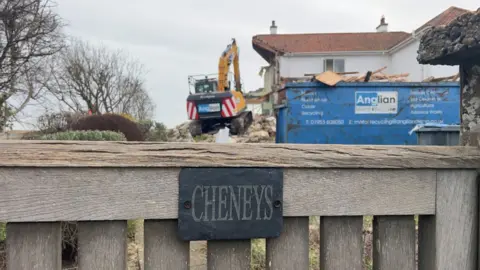 John Fairhall/BBC The Cheneys house. A wooden gate at the forefront of the picture says CHENEYS on a slate sign. In the background is a home being demolished. There is an Anglian Demolition digger and a blue skip.