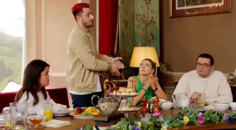 Tom standing at the Traitors castle's breakfast table and gesturing at Alex, who's seated next to him