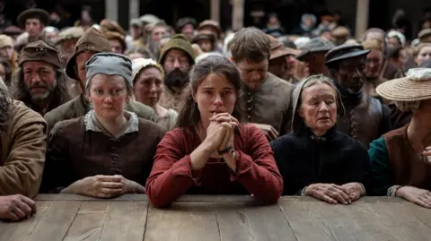 Universal Jessie Buckley in Hamnet, standing with her hands clasped, resting her arms on the stage of a theatre, with the rest of the audience in the background