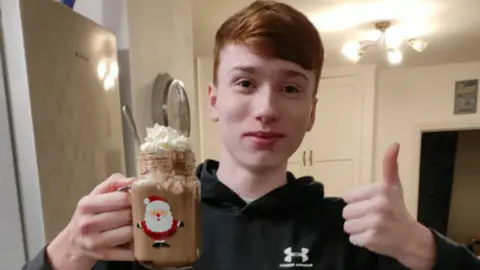 Anita Morris Olly Barnett, a teenager with short ginger hair, wears a black hoody and smiles at the camera. He's holding a hot chocolate in a Santa mug and giving a thumbs-up.