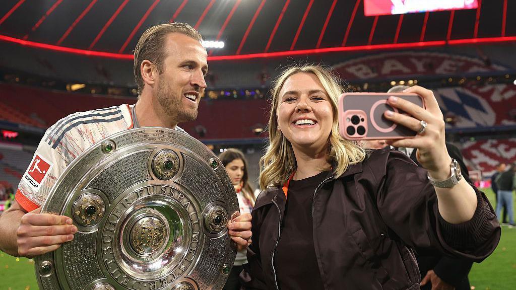 Harry and Kate Kane pose inside a football stadium as Harry holds the Bundesliga shield and Kate takes a picture of them using her pink and grey mobile phone
