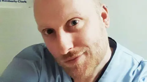 Sheena Millar Close-up of a white blonde man in his 30s, with a light beard and wearing blue scrubs, as he smiles at the camera.