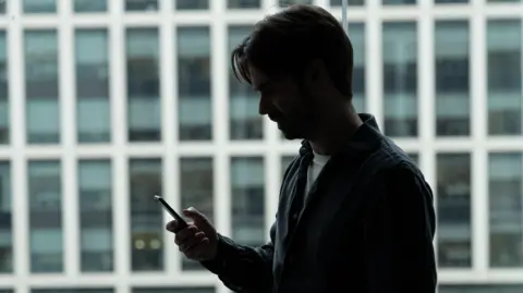 The silhouette of a man holding his phone out to read messages, behind him are rows of office windows.