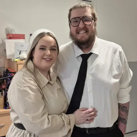 Family handout A blond-haired woman stands next to a man with brown hair and a beard. She has her right hand across his stomach showing a wedding band. The man wears a white shirt and black tie. Both smile at the camera