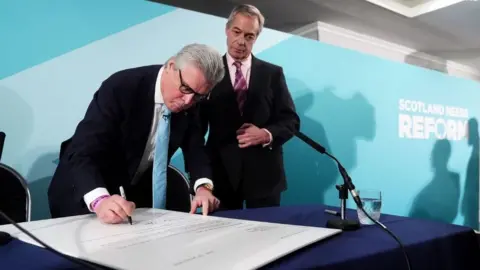 PA Media Malcolm Offord, who has grey hair and glasses, signing a large letter on a blue table. Nigel Farage, who has thinning grey hair, overlooks him. Both are wearing dark suits, and standing in front of a pale blue Reform-branded background.