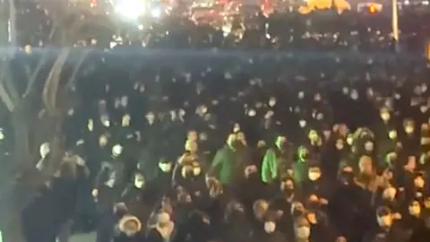 User generated content A large group of people gathered in a street at night. Most wear masks and many hoods. Light streams in, illuminating a tree, with bright lights in the background.