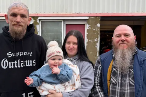 Alex standing in the middle holding Lilah in a cream blanket next to Wesley and Marc in front of a garage. They are all looking at the camera. Wesley, on the left, wears a black hoodie and is bald with tattoos on his head. He has a ginger beard. Alex is wearing a grey hoodie and has long black hair. Baby Lilah is smiling and has one of her hands near her mouth, she has a white bobble hat on and a denim dress. On the right, Marc wears a grey tartan shirt and a blue gilet. He is bald with a long grey beard.