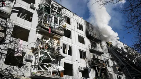 ARIS MESSINIS/AFP via Getty Images A firefighter is standing on a ladder next to a light grey multi-storey building which has white smoke pouring out of it. The twisted remains of balconies can been seen and there is no glass in many of the window frames.