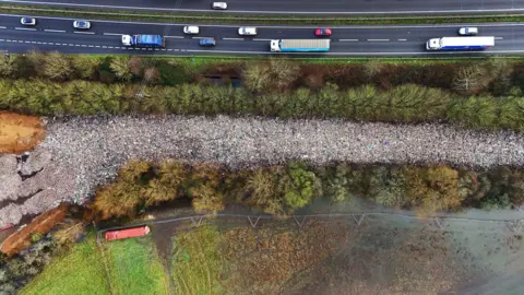 PA Media An aerial "bird's eye" view of a 150m-long stretch of waste dumped in a long line, bordered by trees. On one side lies a busy A-road with cars and lorries. On the other side a flood plain is mostly covered by water.