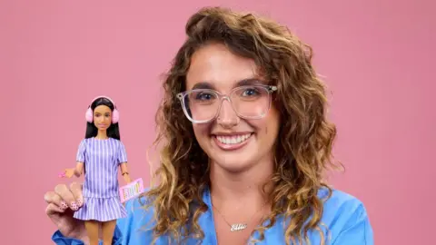 Mattel/PA A woman with wavy light brown / golden hair smiles as she holds a Barbie doll with brunette hair and pink ear defenders. She has glasses on and is wearing a light blue top and is posing against a plain pink background.