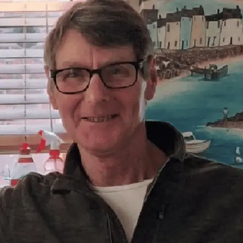 Family Handout A man with brown hair, dark rimmed glasses and a dark jumper smiles into the camera. He is positioned in front of a window with a colourful picture depicting a seaside harbour visible over his shoulder.