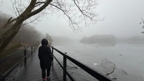 Weather Watchers/Raj's Range A person stands on a wooden bridge wearing a dark parka-style coat. They look out over an icy lake with trees seen amongst the fog in the distance