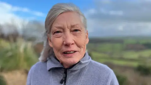 An older woman with shoulder-length grey hair tied back. She wears a grey zip-up fleece and looks at the camera with a neutral expression. The background behind her is out of focus, but she is standing outside with green fields and a cloudy blue sky behind her.