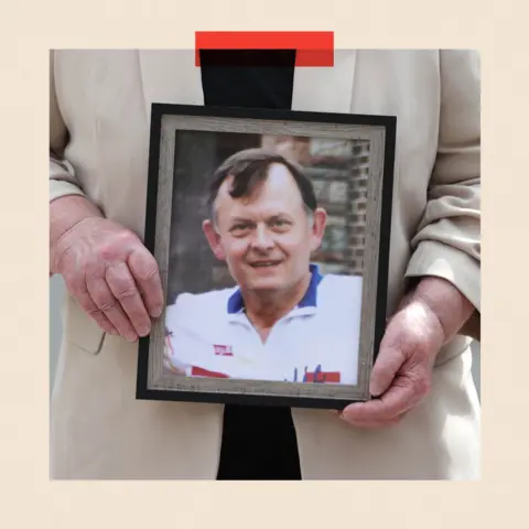 PA Media Cropped image of Bridie Brown, the widow of Sean Brown, holding a picture of him. It is an A4 framed photo and he is wearing a white polo neck t-shirt