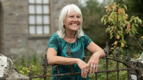 Jon Pountney/Literature Wales A woman in a green blouse stands in a garden, resting her arms against the gate, smiling into the distance. She wears rings on both hands and leaf-shaped earrings.
