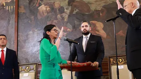 Getty Images Delcy Rodr&iacute;guez wears a green dress and places her left hand on a red velvet covered book as she raises her right hand and is sworn in as interim president