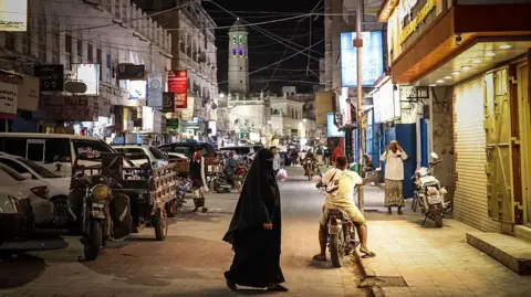 Fadel SENNA / AFP via Getty Images A woman clad in black walks in a street with buildings on both sides , with a man on a motorbike behind her and a mosque minaret in the background