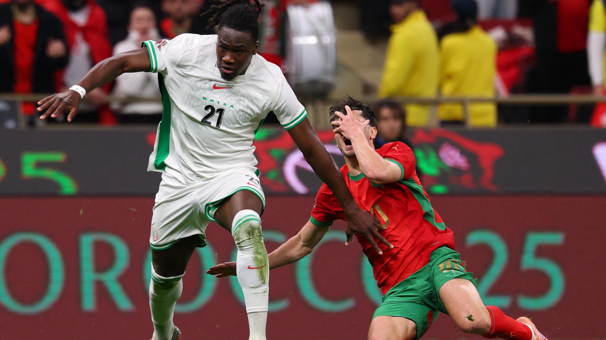Morocco forward Brahim Diaz, in red jersey, green shorts and red socks, goes down clutching his face with his left hand while Nigeria defender Calvin Bassey, all in white, strides away with his arms outstretched. A billboard for Morocco 2025 and some fans are visible in the background