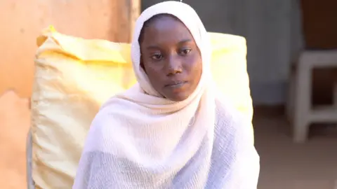 Ikram is sitting on a chair outside with a yellow cushion. She's wearing white robes and a white headscarf.