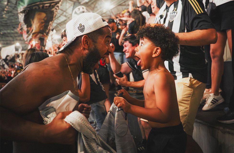 A father and child celebrate Atletico Mineiro scoring. They are both screaming at each other with joy. They are surrounded by cheering supporters in black-and-white striped jerseys.