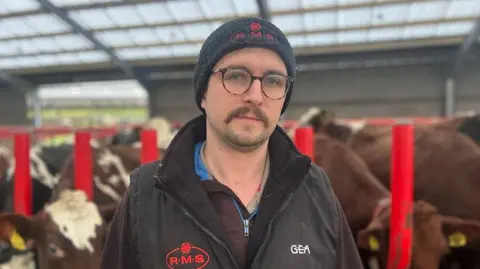 BBC News Farmer Adam Johnstone, wearing a black fleece, gilet and hat, with a glum expression standing in front of his herd of cows in a dairy shed