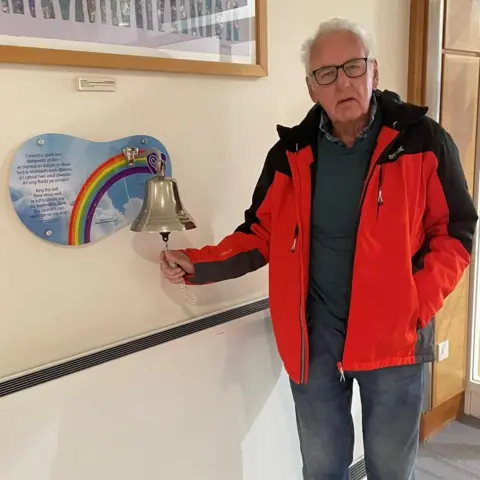 Noel Thomas Noel, wearing a red and black rain coat, ringing a metal bell in a hospital corridor. There is a rainbow mural on the wall the bell is attached to.