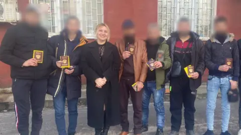 Telegram Polina Azarnykh standing smiling in a long black coat and smart black boots in a line of men wearing jeans, casual shoes and jackets. Their faces have been blurred to conceal their identities. They are holding up Russian travel documents.