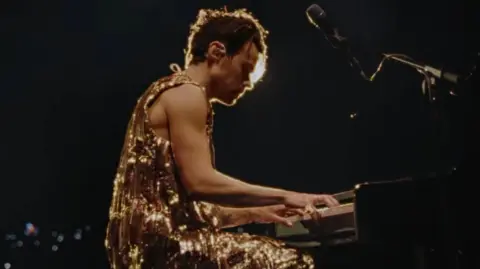 Harry Styles / YouTube Harry Styles sits at a piano in a sparkly gold outfit during the final date of his Love On Tour tour in 2023