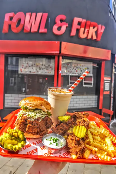 Fowl and Fury Fowl and Fury sign, red letters on a black building. Someone is holding up a tray stacked with a chicken burger, chips, gherkins and a milkshake.
