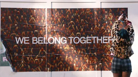 Getty Images A woman holds her phone camera up high above her head to take a photo of a Harry Styles "We Belong Together" poster on a wall in Soho, London.