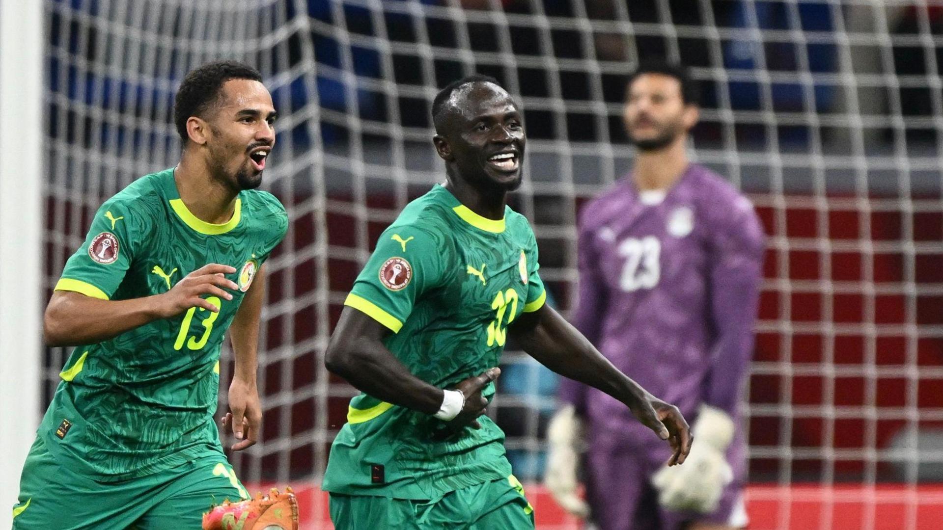 Sadio Mane wheels away in celebration with a smile on his face as he is chased by Senegal team-mate Iliman Ndiaye. Both players wear green Senegal kits, while Egypt goalkeeper Mohamed El Shenawy, wearing purple, is seen dejected in the background standing in front of a white net