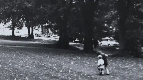 Janet Brocklehurst A black and white photo of two young children holding hands and walking in a park.
