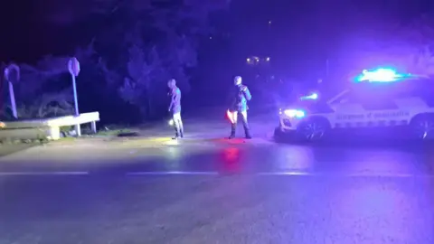 Mossos Two emergency services workers stand in the middle of a road turning next to an emergency response vehicle with its lights on in the dark.