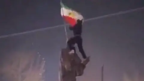 Eyewitness image A man cstands on the top of a statue with the Iranian flag flying. The statue represents a man