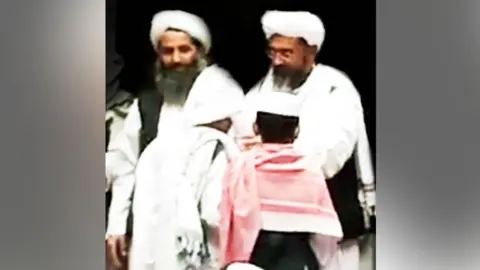 A grainy image showing two men dressed in white with turbans. The man on the left has a large beard which is greying. The man on the right wears glasses, with a slightly shorter beard. In front of them are what appears to be two boys. File photo