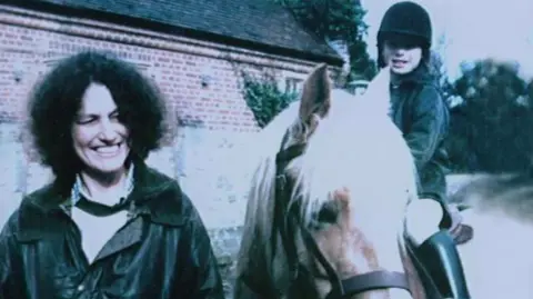 PA Media A woman with shoulder length dark curly hair and wearing a dark coloured jacket and light coloured blouse is smiling as she stands next to a young girl riding a horse. The girl is also smiling. A brick building and trees can be seen behind them.