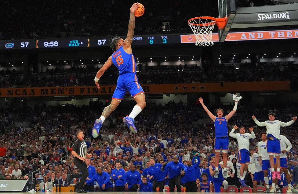 Basketball player Alijah Martin leaps high toward the hoop for a slam dunk during a game. The player wears a blue and orange uniform, and teammates on the bench celebrate in the background as a packed arena watches.