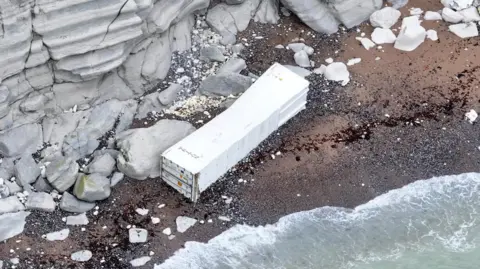 Eddie Mitchell An aerial shot of a white container which is washed up on a small beach which has a rock cliff face behind it.