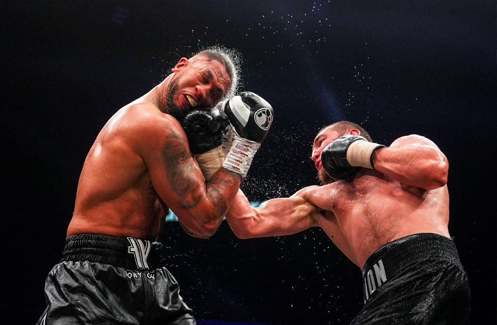 Tony Yoka receives an uppercut punch from Arslan Yellyav during their fight in Paris. Sweat sprays into the air from the impact, and both fighters wear black trunks and gloves.