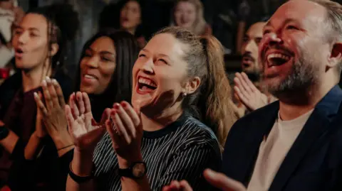 Getty Images A multiracial audience of men and women laughing and applauding while watching a comedy show