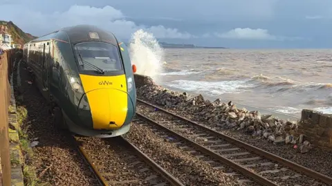 BBC/Johnny Rutherford A GWR train passes a section of crumbled wall. It is next to a churned up sea.