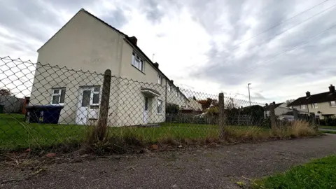 BEN SCHOFIELD/BBC Houses in Sawston. The image has been taken low down - in the foreground is a tarmac footpath, with a small amount of grass verge visible in the bottom right corner. Running alongside the path is a wire mesh fence, with concrete supports every couple of metres. Beyond the fence, we are looking towards the front corner of one of the houses, so that the front door and front windows are facing to the right of the image. There is grass surrounding the house and black wheelie bins against one wall. The houses have cream-coloured walls and the sky above is marked with grey, moody clouds.