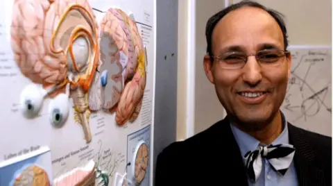 A man with glasses and a bow tie stands smiling beside a display showing an illustration of a human brain