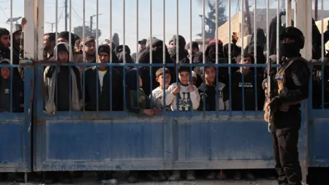 EPA Syrian security forces stand in front of the gate of the al-Hol camp, behind which stand family members of suspected IS members, in Hassakeh province, Syria (21 January 2026)