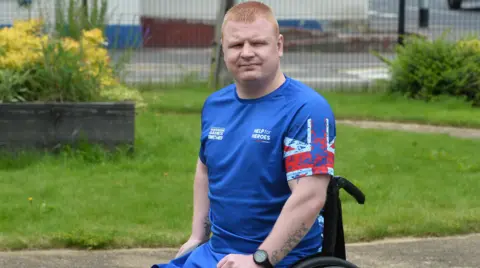 Pacemaker Andy Allen in his wheelchair. He is wearing a blue t-shirt and has ginger hair. He is in an outdoor area with some grass visible behind him, and he is looking directly at the camera with a neutral expression on his face.