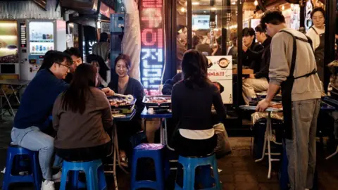 Getty Images People eat barbecue on tables and stools outside a restaurant.