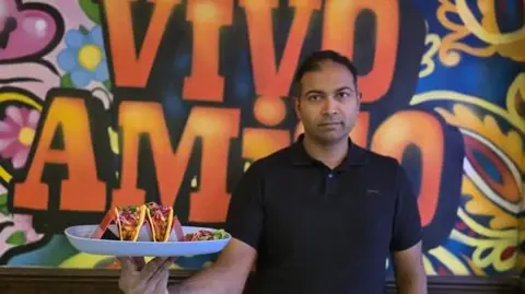 Rajendra Vikram Kupperi Rajendra Vikram Kupperi wearing a black short-sleeved shirt is holding a large white plate with three vibrant tacos. Each taco is filled with colorful ingredients, including shredded lettuce, diced tomatoes, and possibly meat, topped with bright pink pickled onions. The background features a bold, colorful mural with swirling patterns and large orange letters spelling &ldquo;VIVO AMIGO&rdquo; against a mix of blue, yellow, and pink designs.