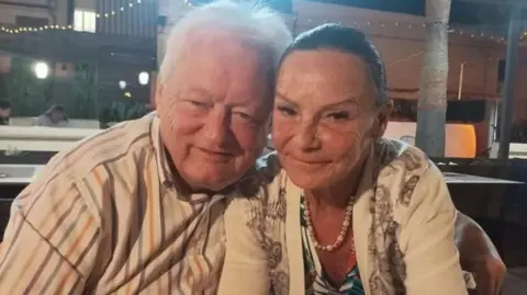 Supplied Bill Trotter is a man of 73 who is wearing a striped shirt and has grey hair. He is smiling and leaning into his wife next to him who is a similar age and has dark hair tied back and is wearing a blue top and a white cardigan. They are sitting close together, smiling gently.